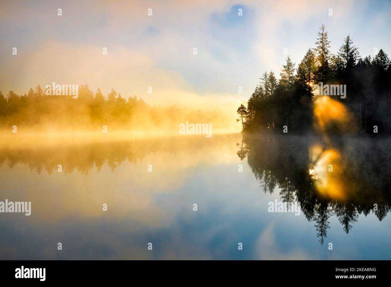 Sonnenaufgang im Gegenlicht mit Nebelschwaden über dem spiegelglatten Moorsee Étang de la Gruère im Kanton Jura, Schweiz Foto Stock