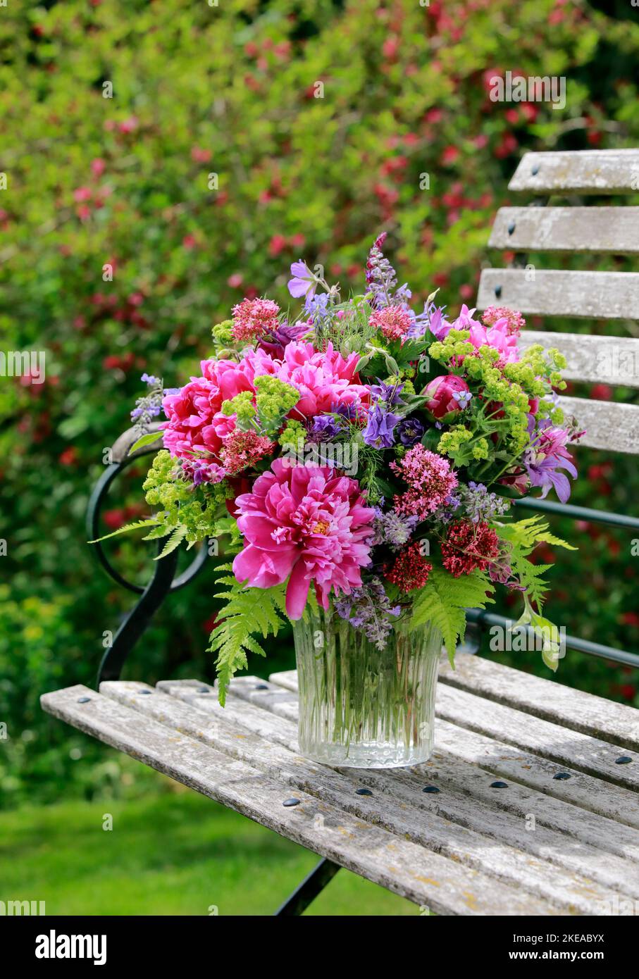 Bungter Blumenstrauss in rot, pink und violett Farbtönen mit Pfingstrosen und Akeleien, steht in Glas-Vase auf dekorativen Holz-Gartenbank Foto Stock