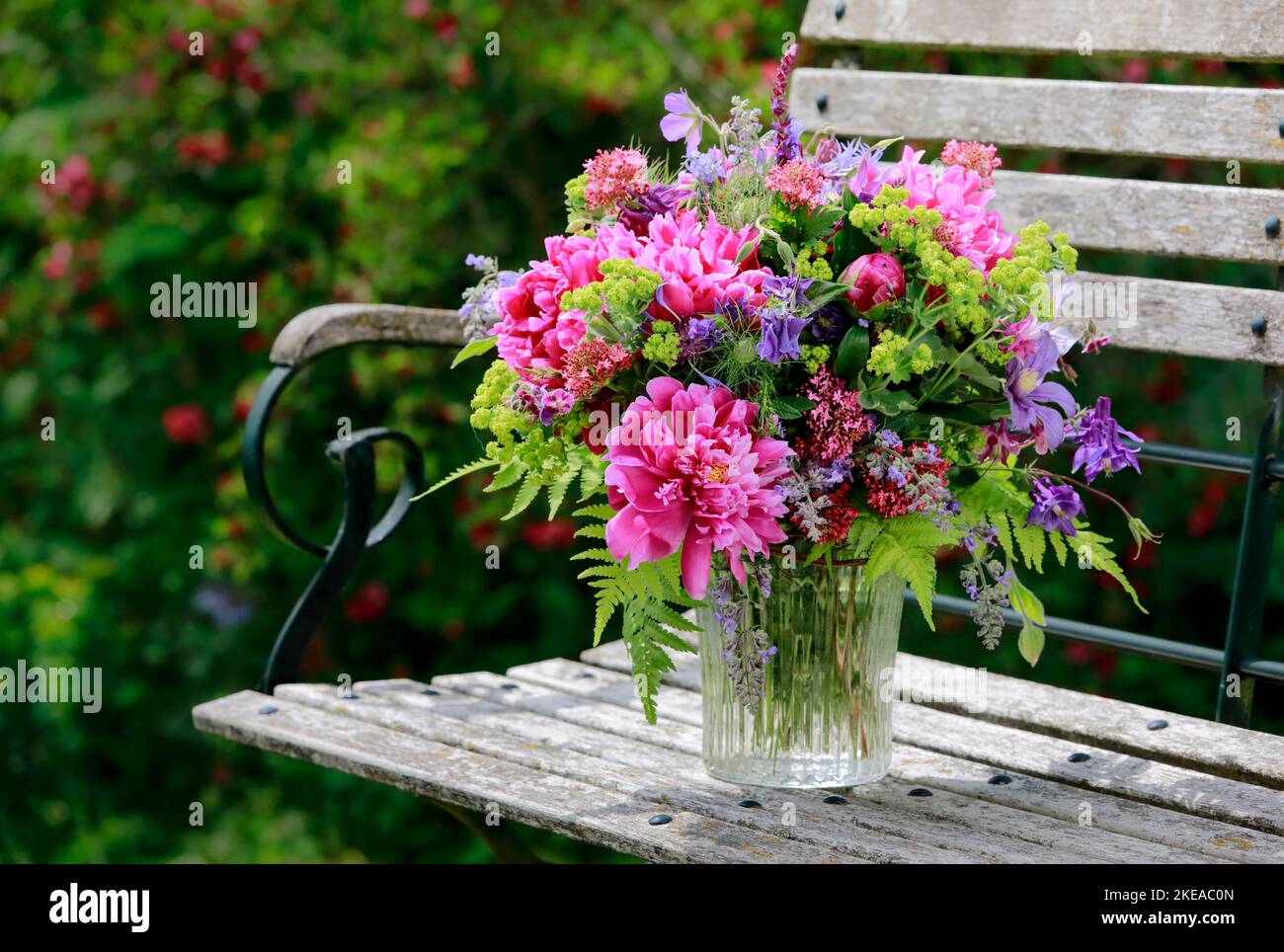 Bungter Blumenstrauss in rot, pink und violett Farbtönen mit Pfingstrosen und Akeleien, steht in Glas-Vase auf dekorativen Holz-Gartenbank Foto Stock