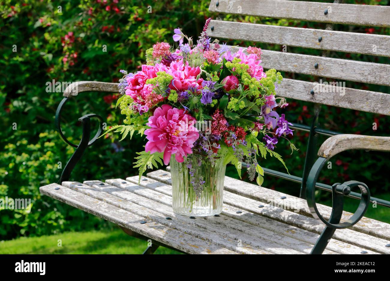 Bungter Blumenstrauss in rot, pink und violett Farbtönen mit Pfingstrosen und Akeleien, steht in Glas-Vase auf dekorativen Holz-Gartenbank Foto Stock