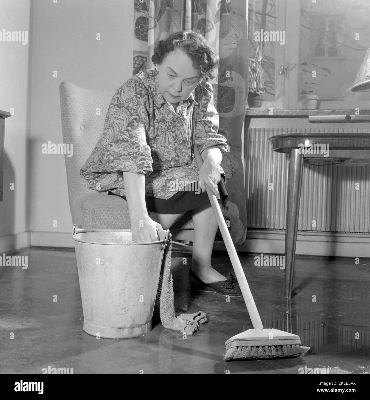 Pulire il pianale sul retro. Una donna sembra molto stanca e logora quando si siede su una sedia, avendo un riposo nel lavoro di pulizia dei pavimenti. Svezia 1956 Conard Ref 3160 Foto Stock