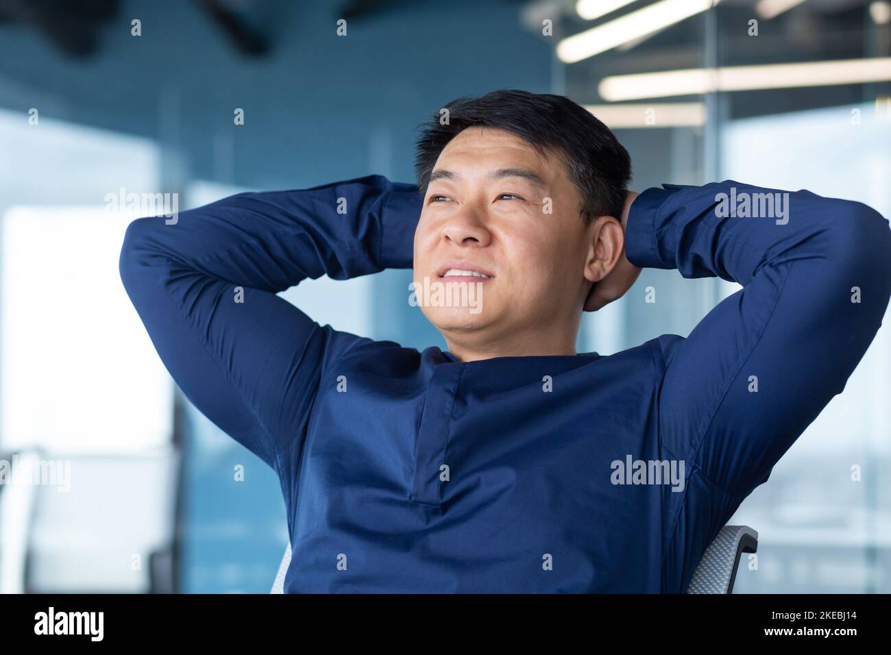 Uomo d'affari felice che riposa dopo il lavoro completato con successo uomo asiatico sorridente e felice guardando finestra pensando e sognando sul progetto futuro, uomo in camicia con le mani dietro la testa all'interno dell'ufficio. Foto Stock