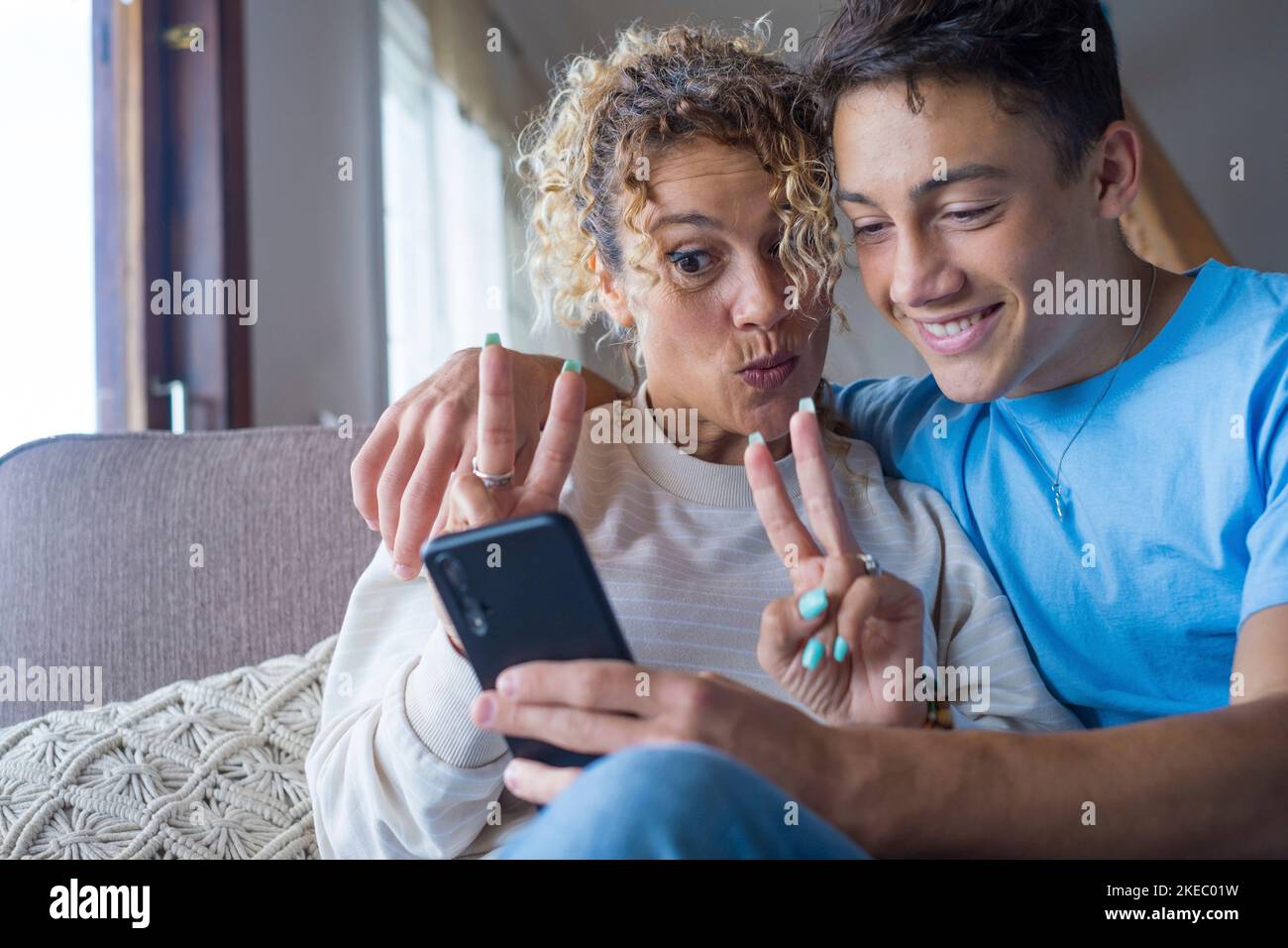 Sorridente riposo madre di mezza età di 40s anni con un figlio adulto che utilizza lo smartphone insieme, felice giovane uomo godersi il fine settimana in famiglia con la mamma prendere un selfie, divertirsi a casa Foto Stock