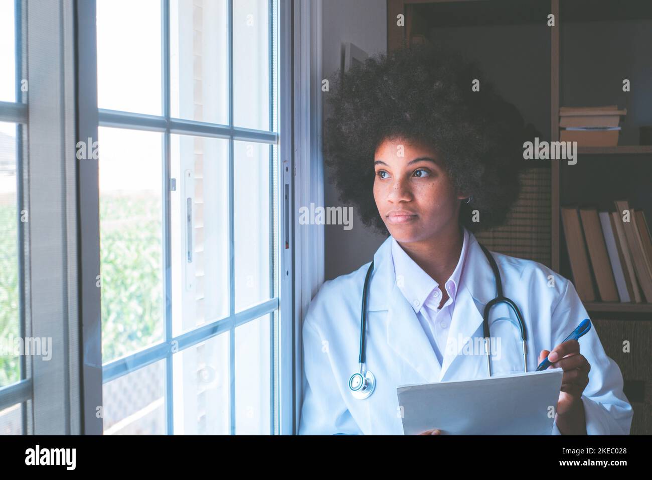 Medico afro-americano premuroso con stetoscopio, documenti e penna che guarda fuori dalla finestra della sua clinica. Serio lavoratore di heathcare di fronte contemplativo ammirando la vista dalla finestra Foto Stock