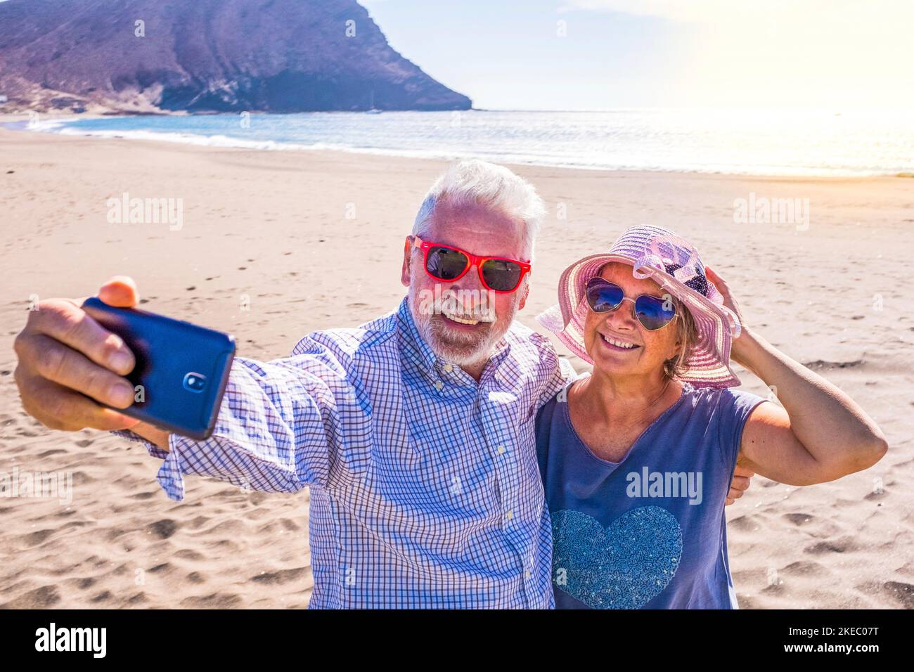 coppia di persone mature o pensionati che si godono le loro vacanze e il tempo estivo insieme sulla sabbia della spiaggia con il mare o l'oceano sullo sfondo - due anziani pensionati prendere un selfie guardando il telefono Foto Stock