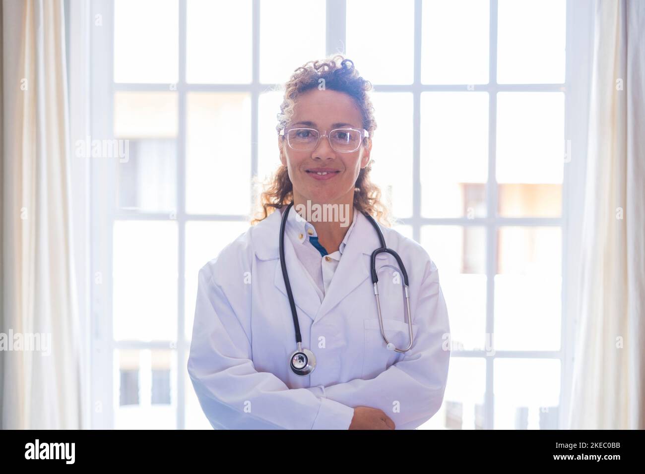 Portriat della giovane dottoressa in uniforme e stetoscopio in piedi contro la finestra in clinica. Pratico medico di prima linea o operatore sanitario sicuro negli occhiali in ospedale. Foto Stock