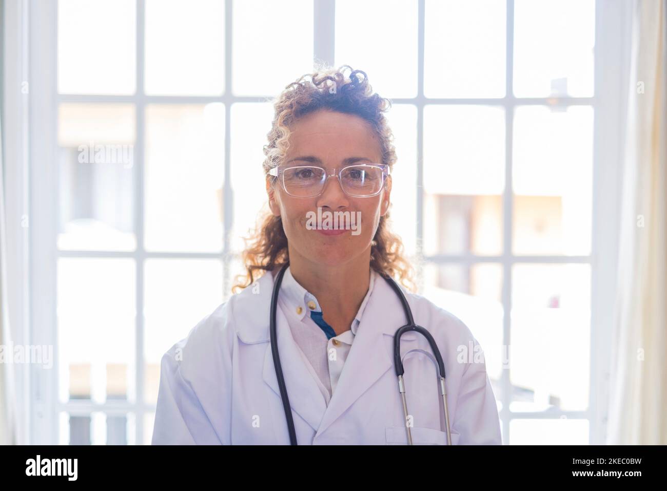 Portriat della giovane dottoressa in uniforme e stetoscopio in piedi contro la finestra in clinica. Pratico medico di prima linea o operatore sanitario sicuro negli occhiali in ospedale. Foto Stock