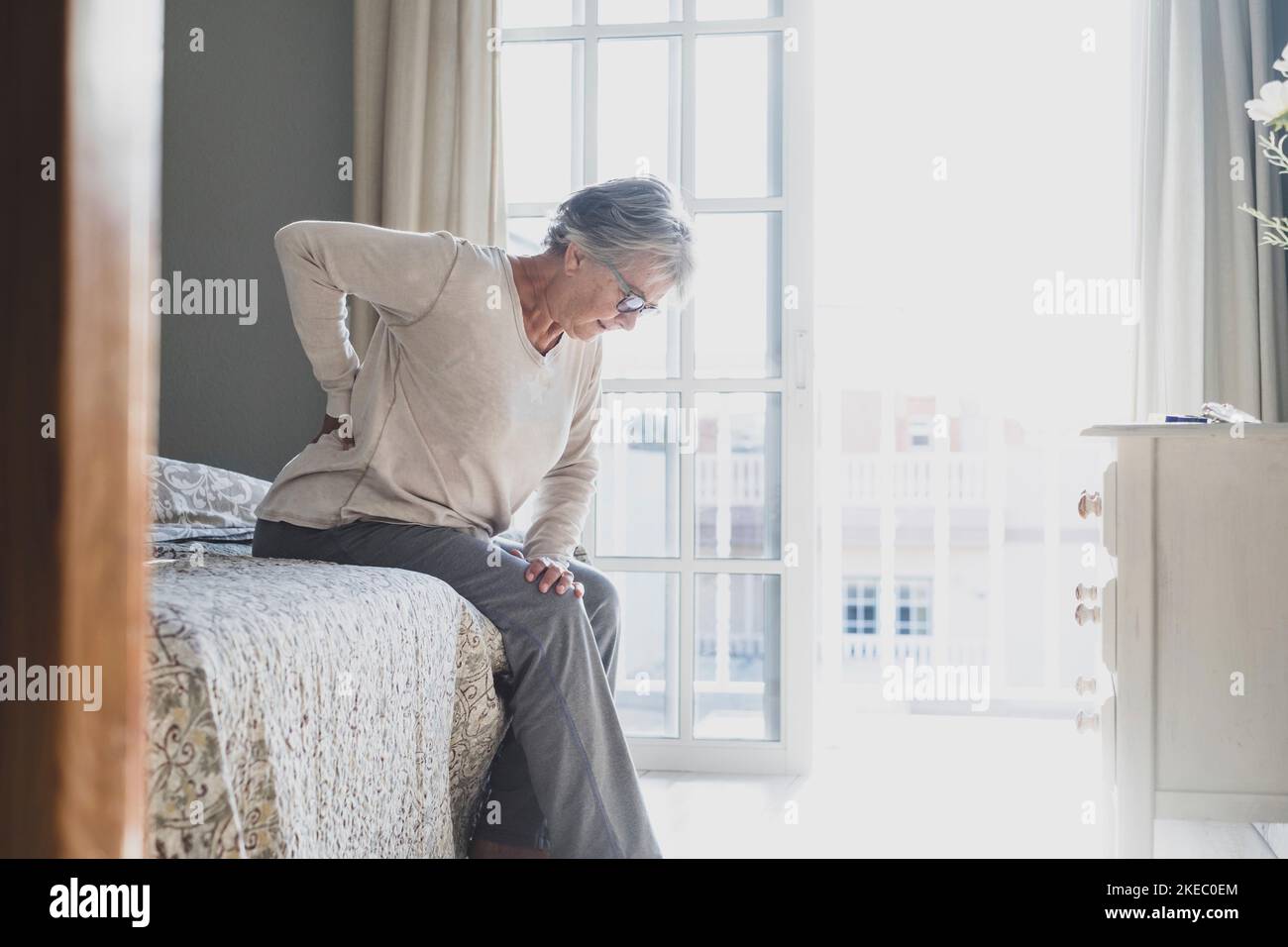 Donna anziana che soffre di mal di schiena dopo il sonno, strofinando i muscoli rigidi, vecchia donna seduta sul letto toccare la parte inferiore della schiena sensazione di disagio a causa di letto a disagio a casa Foto Stock