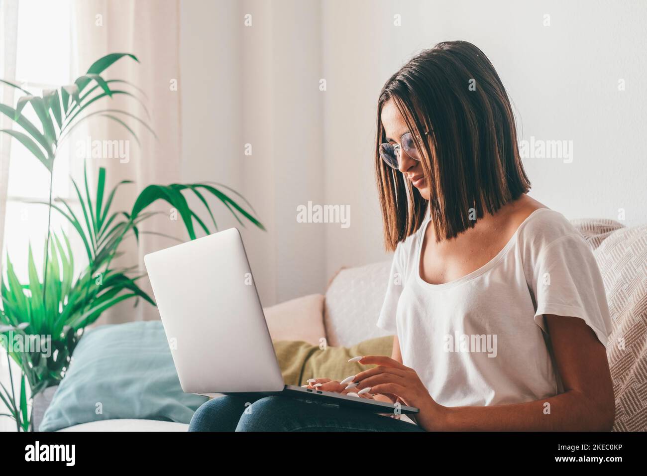 Giovane donna felice negli occhiali che scrivono usando il laptop mentre si siede sul divano nel soggiorno della sua casa. Bella donna che trascorre il tempo libero utilizzando il computer portatile. Freelance che lavora da casa Foto Stock