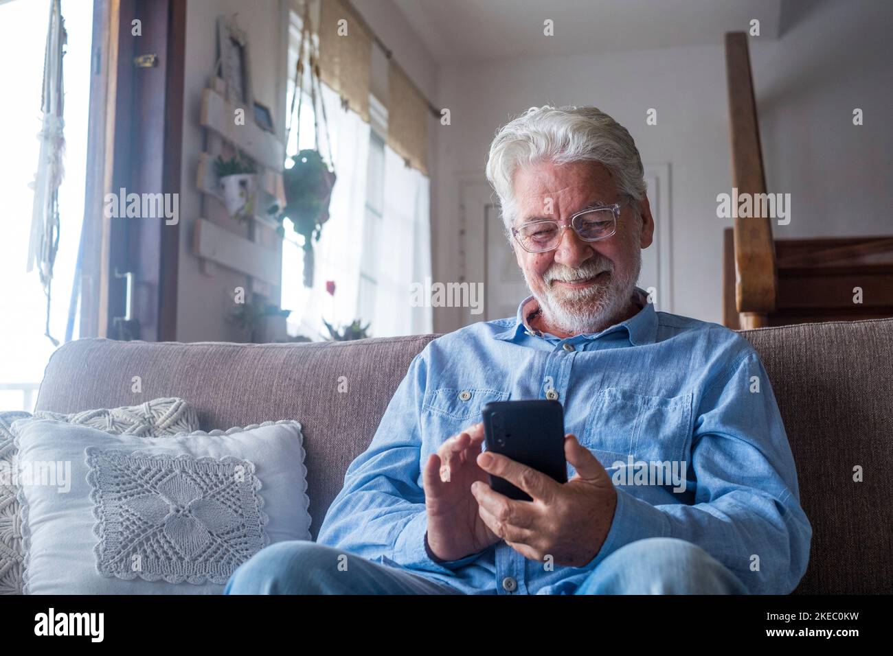 Il vecchio sorridente seduto sul divano nel salotto che tiene il telefono, godendo usando lo smartphone sentendosi soddisfatto inviando i messaggi, denominando gli amici, surfing il concetto in linea di fotoricettore Foto Stock