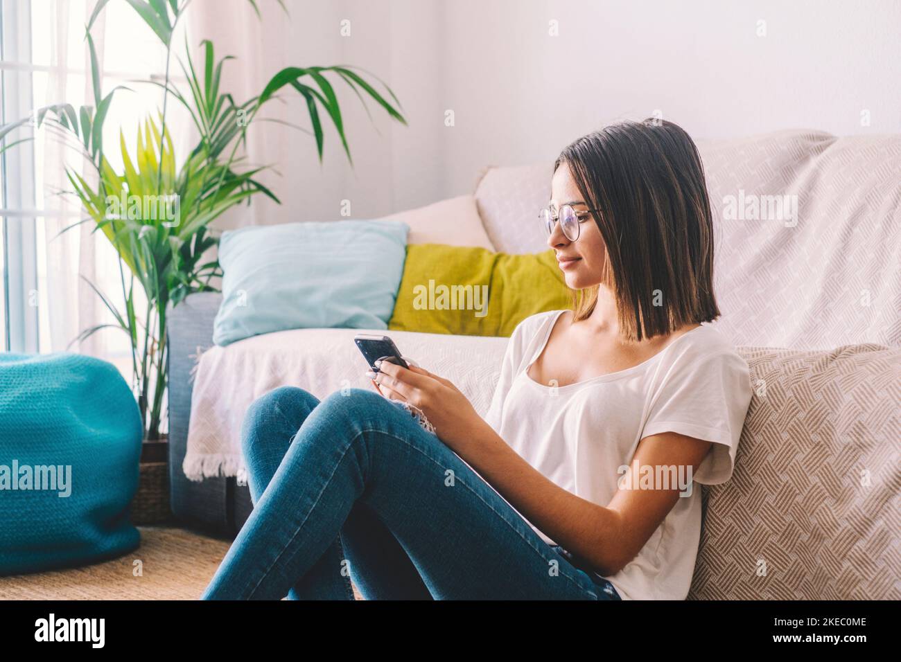 Donna seria nella messaggistica di testo degli occhiali che utilizza il telefono cellulare mentre si siede sul pavimento nel soggiorno della sua casa. Bella donna che trascorre il tempo libero utilizzando lo smartphone a casa Foto Stock