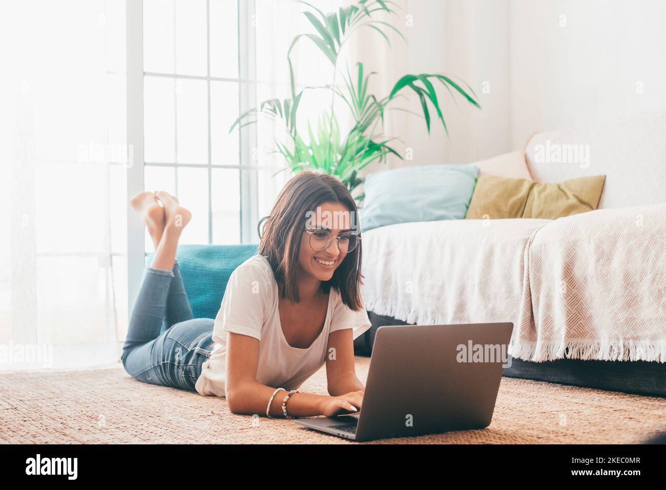 Giovane donna felice negli occhiali che scrivono usando il laptop mentre sdraiata sul tappeto nel soggiorno della sua casa. Bella donna che trascorre il tempo libero utilizzando il computer portatile. Freelance che lavora da casa Foto Stock