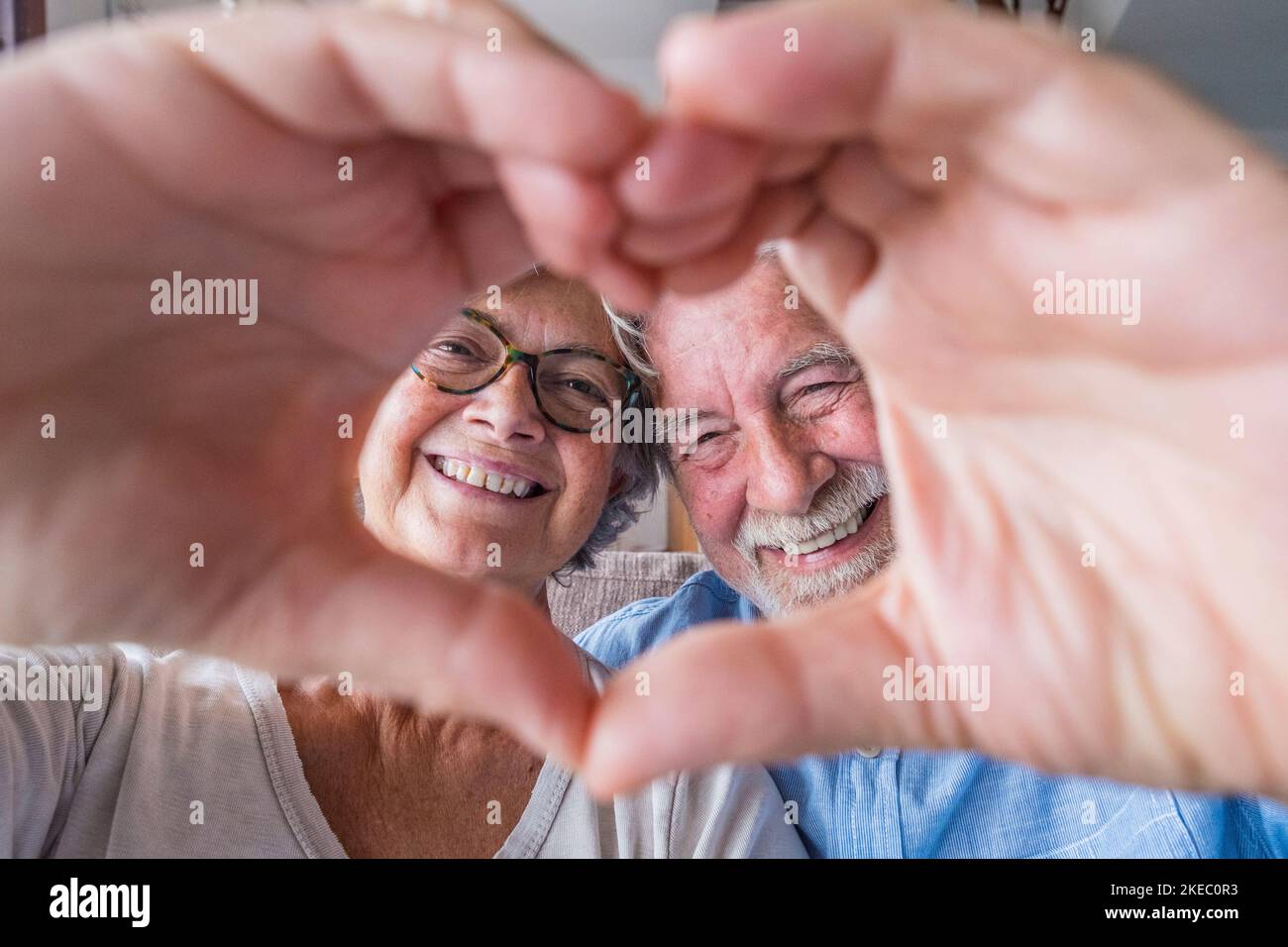 Primo piano ritratto felice sincero anziano di mezza età pensionato coppia di famiglia facendo gesto cardiaco con le dita, mostrando l'amore o dimostrando sentimenti sinceri insieme in interni, guardando la fotocamera. Foto Stock