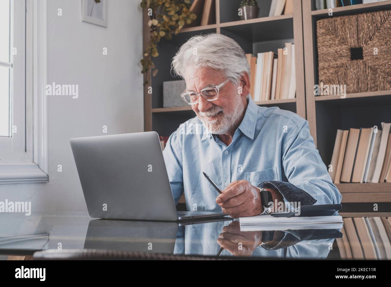 Buon vecchio uomo d'affari senior guardando lo schermo del notebook e sorridendo sulla lettura e-mail seduto a casa ufficio. Uomo anziano che lavora online con la calcolatrice sul tavolo davanti alla libreria sul posto di lavoro Foto Stock