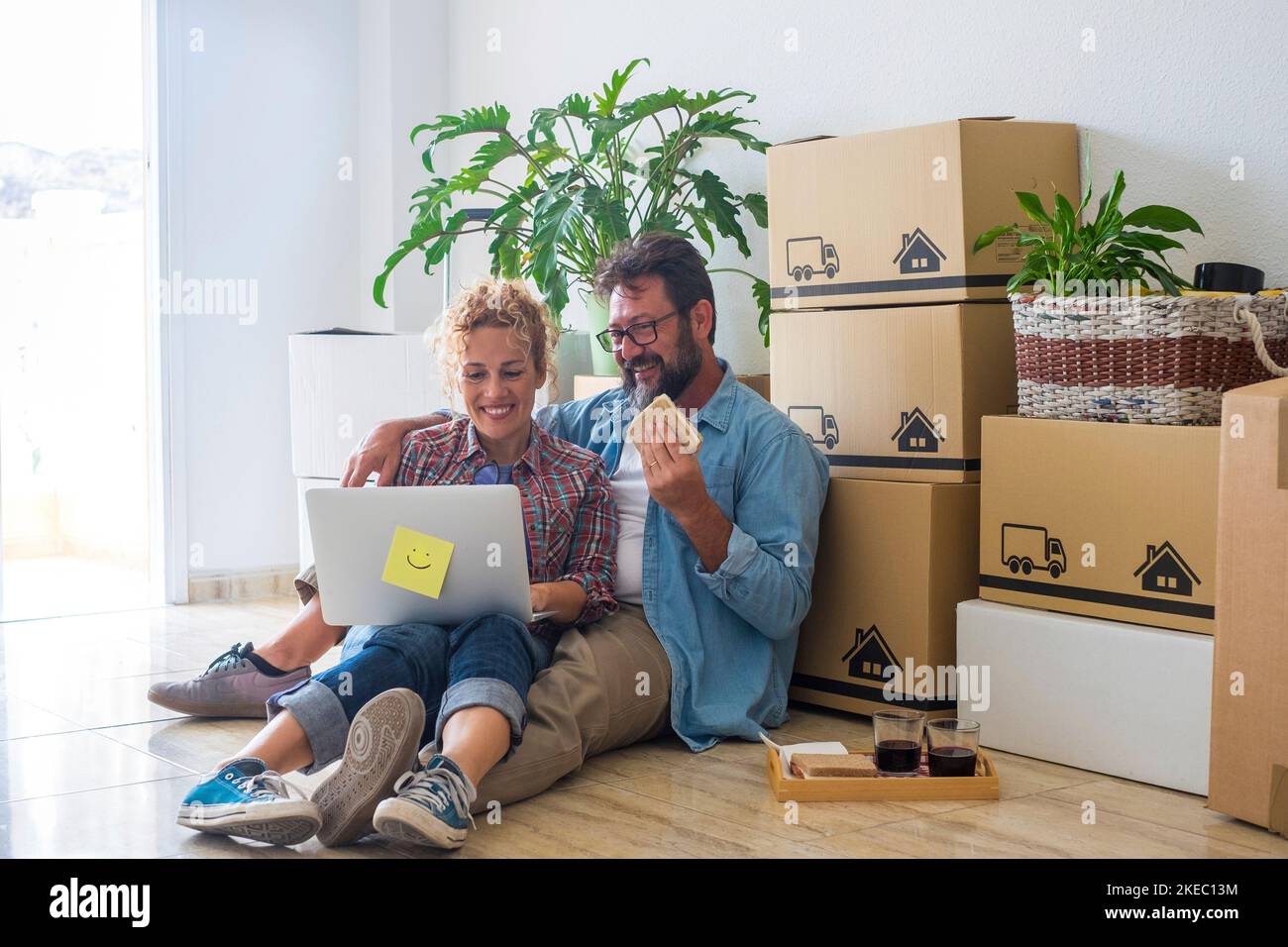 coppia di due adulti e persone felici dopo aver acquistato una nuova casa o appartamento insieme per vivere insieme - due persone a terra utilizzando un computer portatile con scatole e pacchi sulla schiena Foto Stock