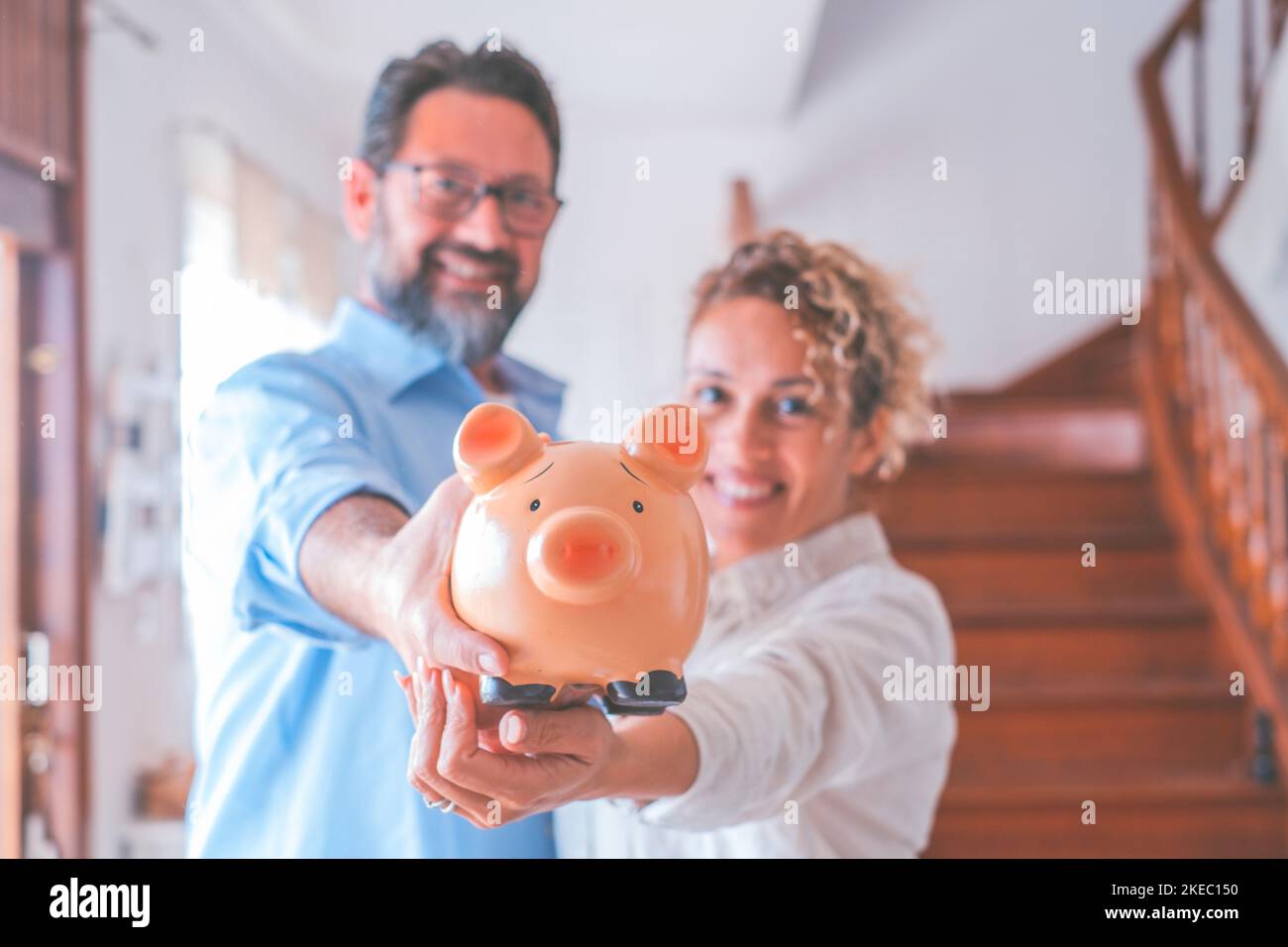 Ritratto di felice coppia caucasica tenendo banca piggy per risparmiare denaro per rendere i loro sogni futuri diventare realtà. Marito e moglie che detengono una banca di porcellini per risparmiare a casa mentre guardano la fotocamera Foto Stock