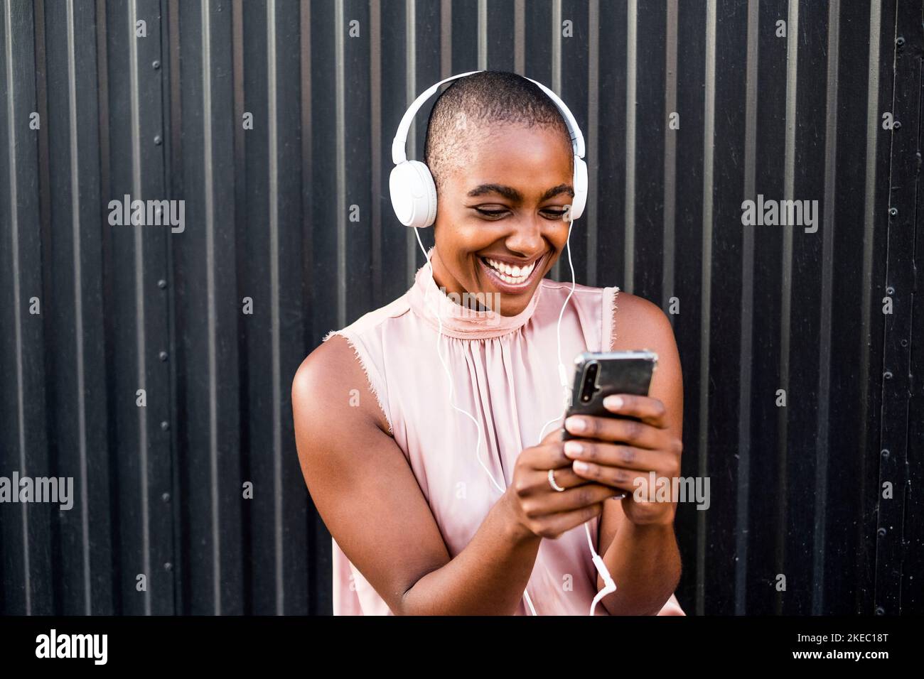 primo piano e ritratto di una giovane donna afroamericana che usa il telefono o la cella mentre ascolta musica con le cuffie - gente felice che si diverte e si diverte a sentire la musica - parete nera sullo sfondo Foto Stock