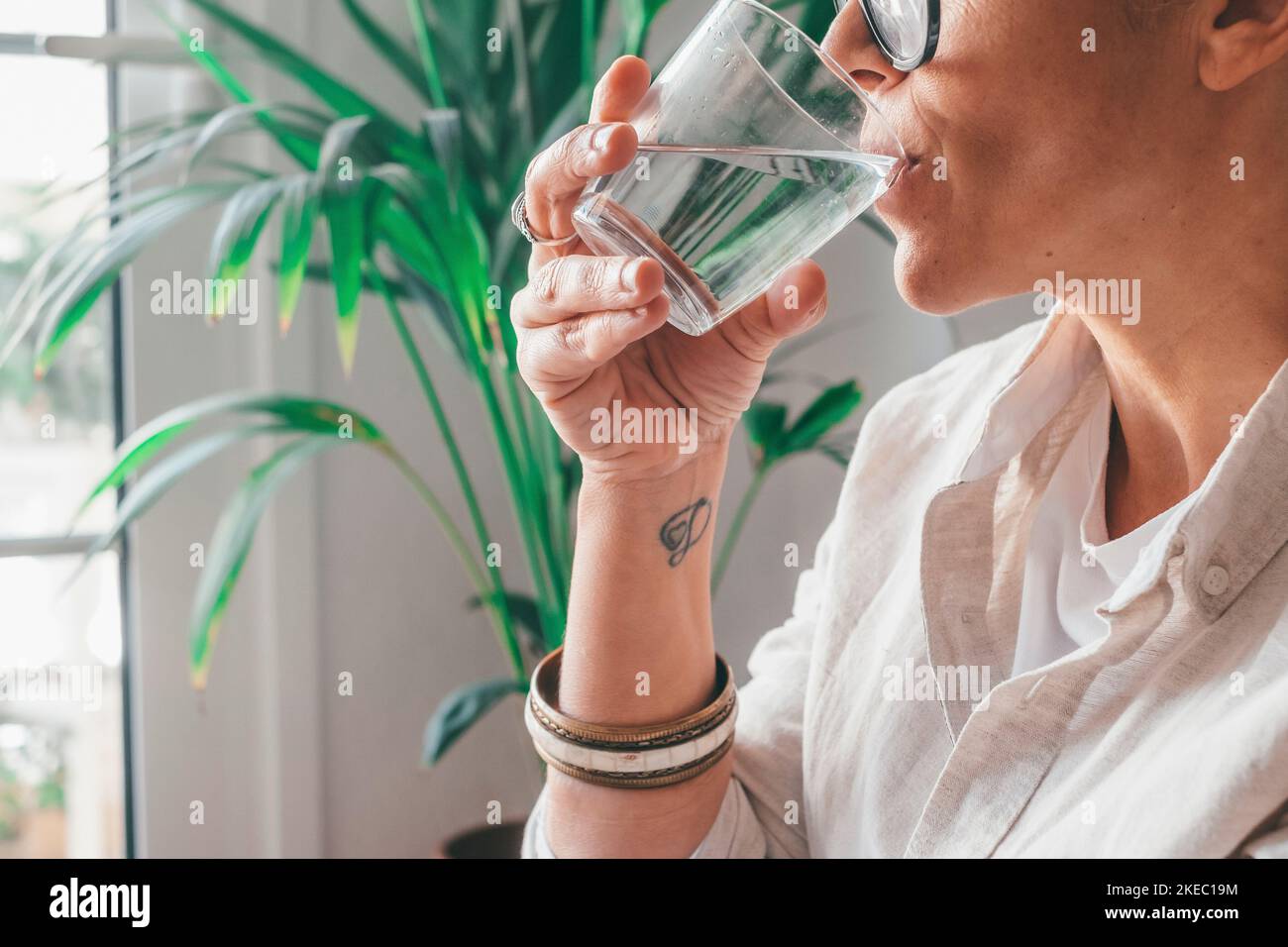 Primo piano di una giovane donna che beve un bicchiere d'acqua fresca nella stanza domestica di casa. Primo piano ritagliato di caucasica thisrty femmina in possesso di vetro trasparente bevande acqua purificata a casa Foto Stock