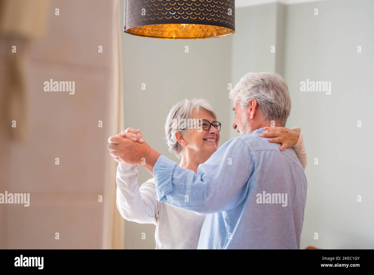 Coppia anziana amorevole romantica che tiene le mani godendo ballare insieme nel soggiorno di casa, coppia anziana felice che festeggia facendo la danza a casa. Vecchio marito e moglie che si divertono in un appartamento moderno Foto Stock