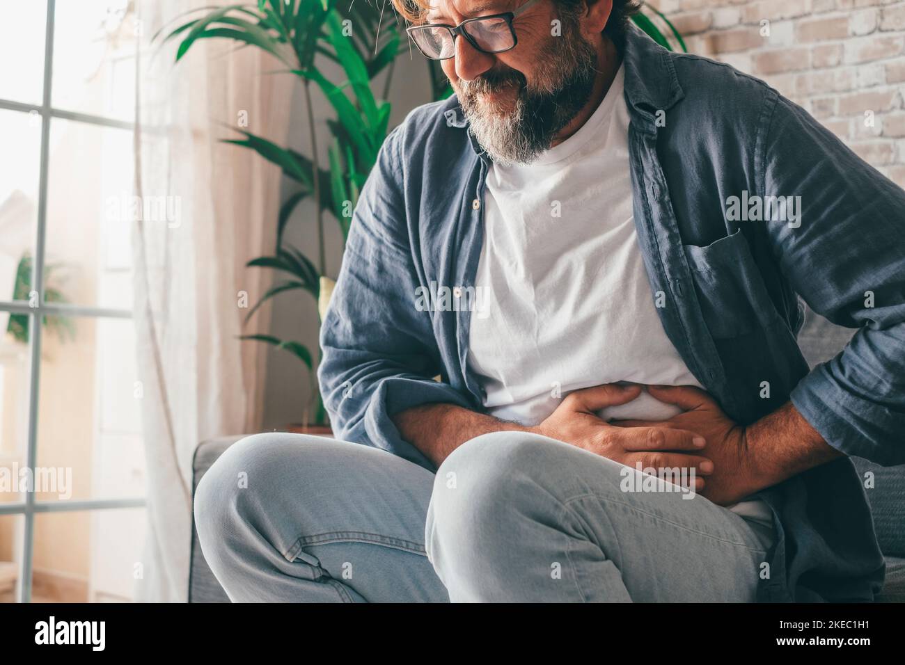 Uomo che soffre con grave dolore allo stomaco seduto a casa. Mano del tipo maturo che tiene l'addome che soffre dal dolore, dalla diarrea o dal problema indigestive. Tipo caucasico che preme sul ventre su sensazione dolorosa Foto Stock