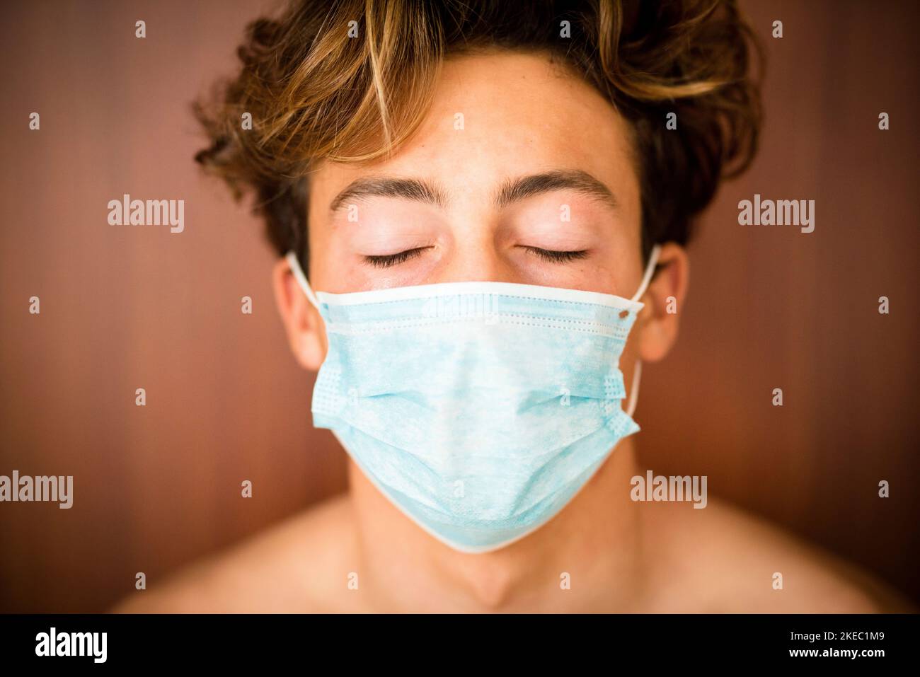 ritratto e primo piano di uomo adolescente o ragazzo millenario con gli occhi vicini che indossano la maschera medica e chirurgica su di lui faccia per prevenire il coronavirus o covid-19 o qualsiasi tipo di malattia o influenza Foto Stock