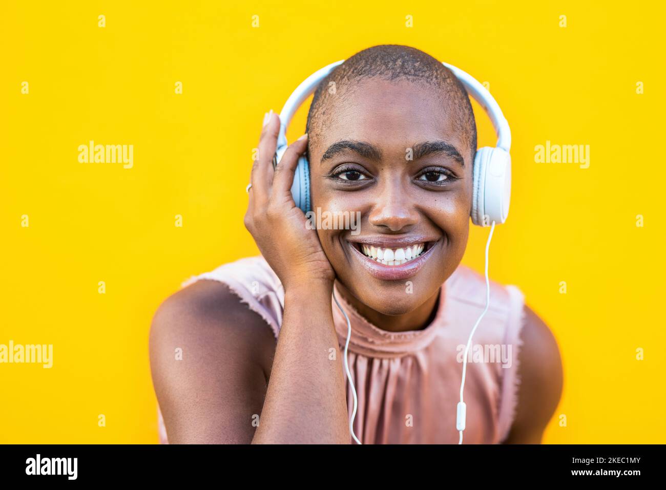 primo piano e ritratto di una giovane donna afro-americana guardando la fotocamera sorridendo e ascoltando la musica con lei cuffie - sfondo giallo Foto Stock