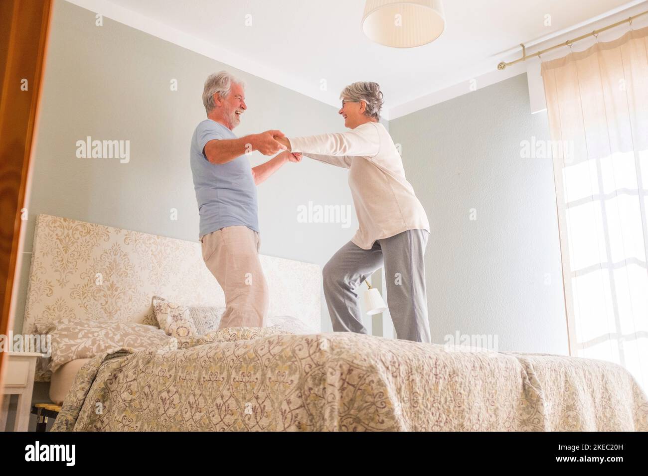 Coppia anziana felice tenendo le mani e ballando insieme a letto a casa. Coppia eterosessuale anziana spensierata e attiva che tiene le mani e balla insieme a letto a casa Foto Stock