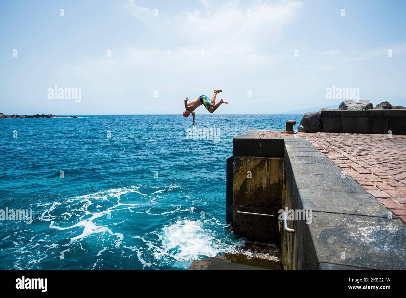 un giovane o un adolescente che si gode il tempo estivo e. vacanze all'aperto in spiaggia saltando di una scogliera a. l'acqua che fa un frontflip - persone attive che si divertono in viaggio Foto Stock