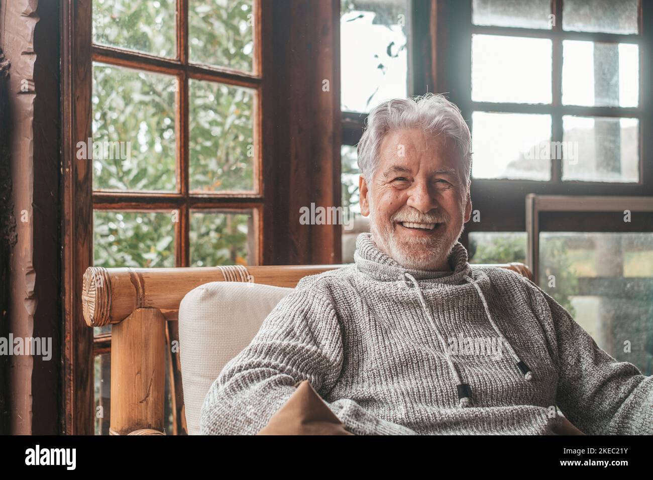 Ritratto di vecchio sorridente e guardando la macchina fotografica seduta sul divano da casa. Closeup uomo persona senior allegro al coperto. Foto Stock