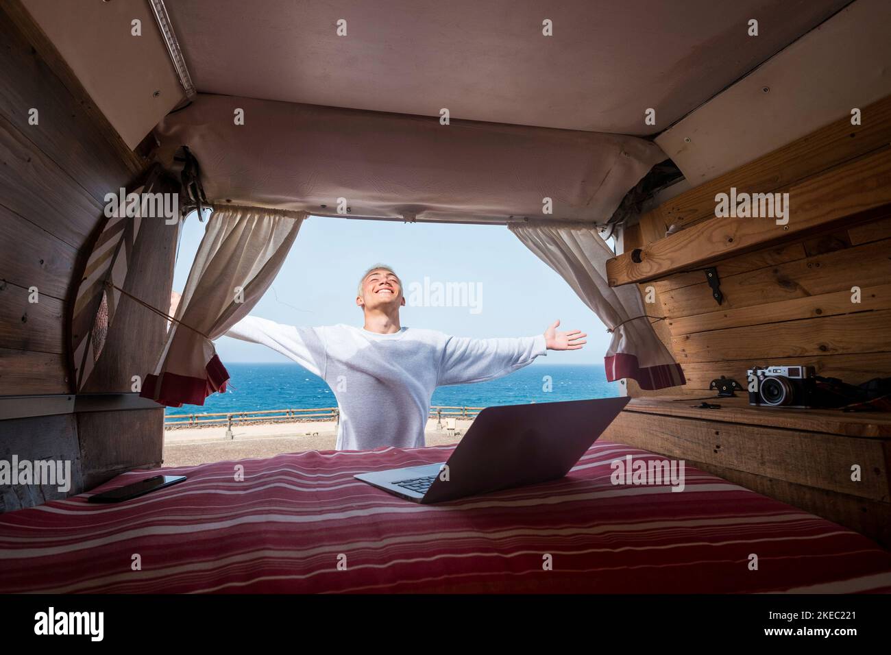 un giovane che si diverte a vivere all'aperto viaggiando e divertendosi da solo con il suo minivan - nomade tecnologia stile di vita - utilizzando e lavorare con il computer portatile con la spiaggia e il mare sullo sfondo Foto Stock