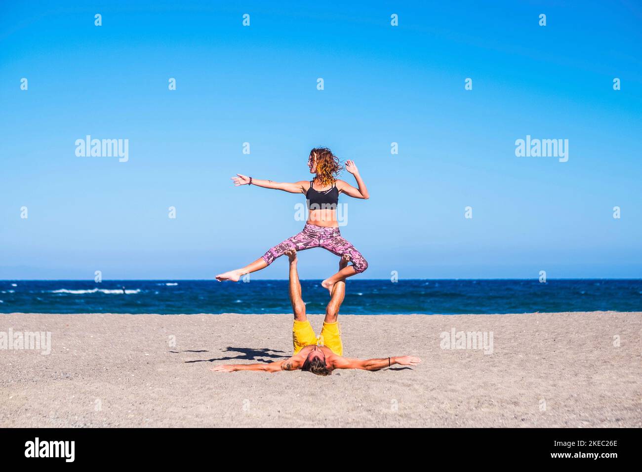 coppia di adulti che fanno l'esercitazione alla spiaggia togetehr sopra la sabbia - due persone che fanno yoga o acro yoga con il mare sullo sfondo - l'uomo che lo holdining ragazza con le gambe Foto Stock