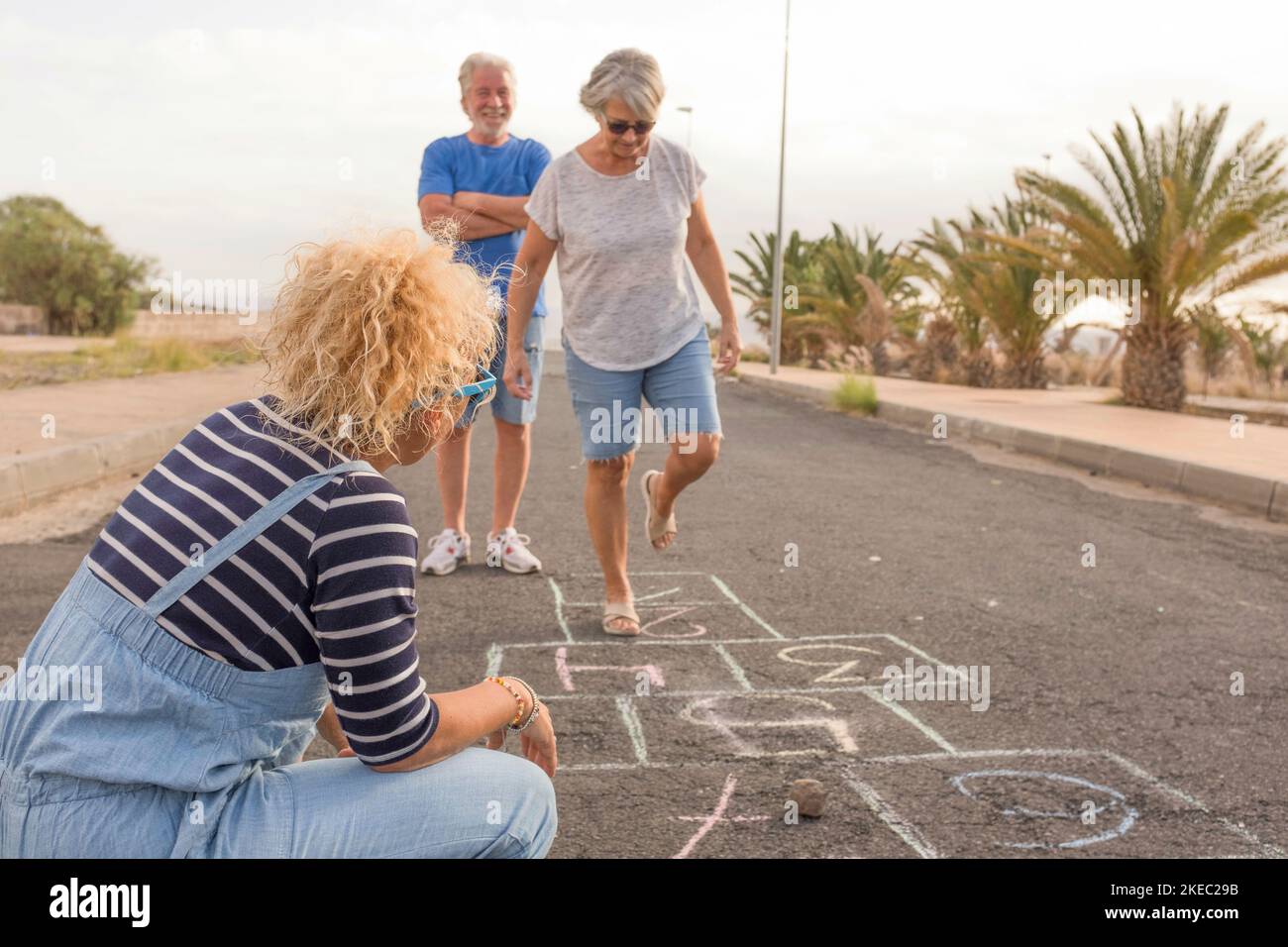 gruppo di tre persone come adulti e anziani - due gli anziani che giocano al hopscotch con una donna curly che guarda la donna matura che salta Foto Stock