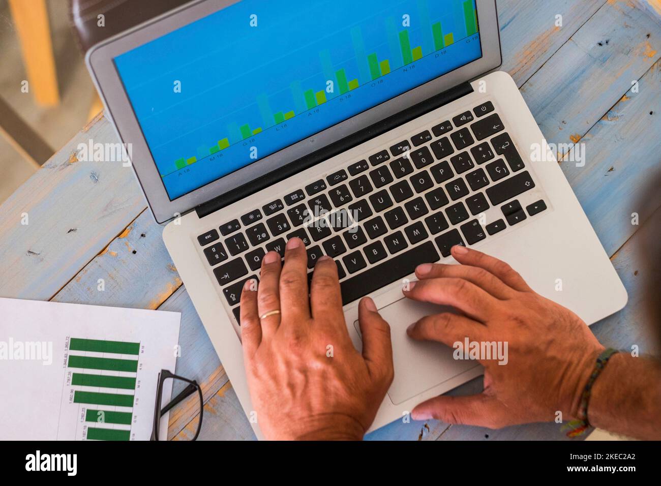primo piano delle mani di un uomo che scrive sul suo computer portatile e guarda alcune statiche del suo lavoro - un tavolo di legno blu Foto Stock