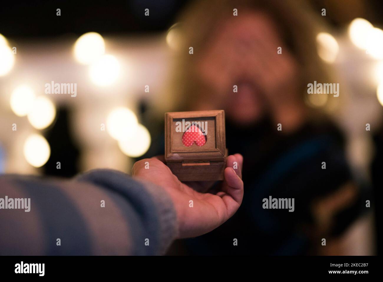 primo piano e ritratto della mano e del braccio dell'uomo chiedere il matrimonio alla sua futura moglie - l'uomo che dà un anello per ottenere allegro alla sua ragazza mentre lei sta coprendo i suoi occhi con le mani Foto Stock