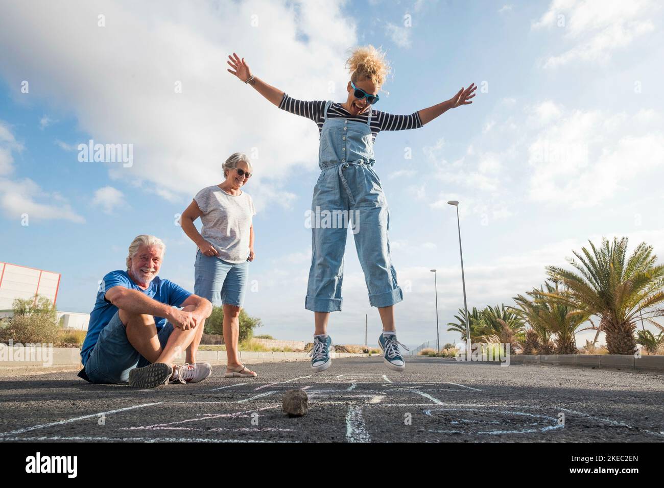 gruppo di tre persone caucasiche che si divertono e giocano il hopscotch insieme - bella donna che salta nel mezzo dell'asfalto e due anziani che la guardano Foto Stock