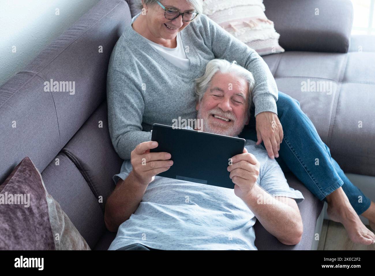 coppia di due anziani o persone mature a casa avendo divertimento insieme guardando lo stesso tablet - pensionato sdraiato sul divano con la moglie seduta Foto Stock