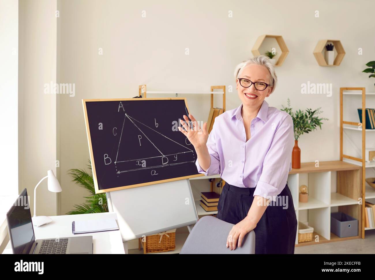 Un insegnante adulto con pelo grigio positivo si trova accanto al tavolo con laptop e lavagna Foto Stock