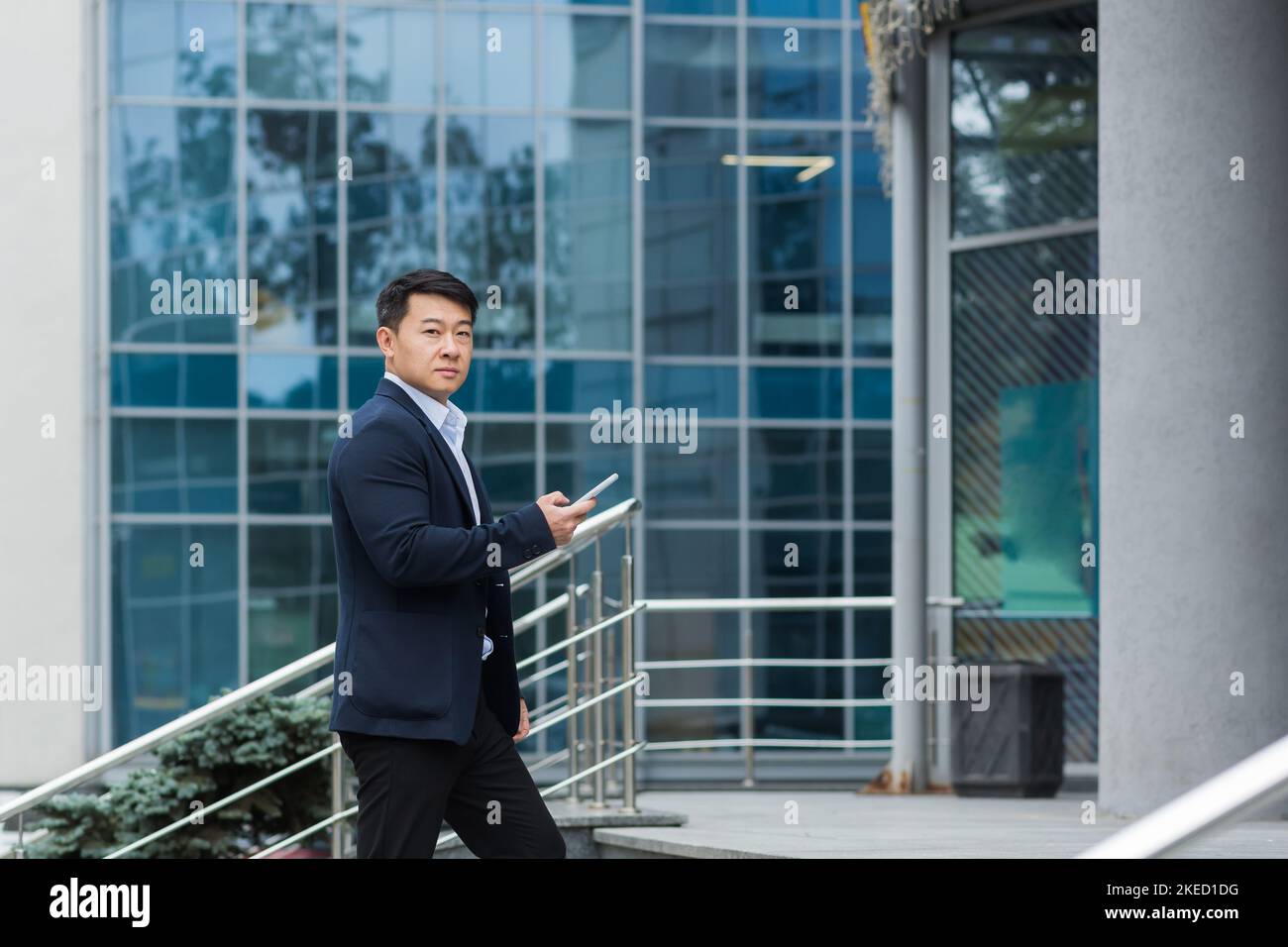 Un giovane bell'uomo asiatico va a una riunione, un'intervista per un lavoro in un centro ufficio. Sta in piedi sulle scale con un telefono e documenti in mano, guardando la macchina fotografica. Foto Stock