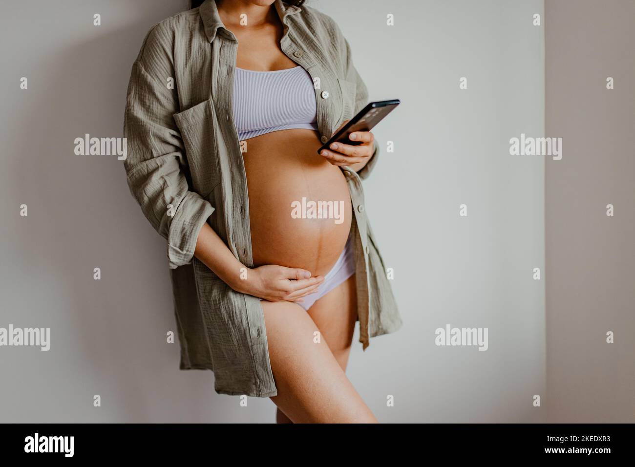 Donna incinta che usa l'app per telefono per la lezione prenatale di gravidanza online o la lettura sulla maternità indossando biancheria intima e camicia casual in cotone a casa Foto Stock