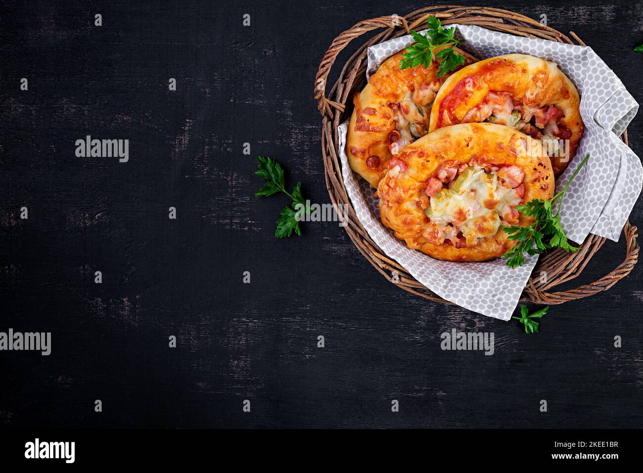 Mini pizza per bambini, piccole torte aperte con prosciutto, salsa di pomodoro, mozzarella e formaggio in cestino su fondo scuro. Vista dall'alto Foto Stock