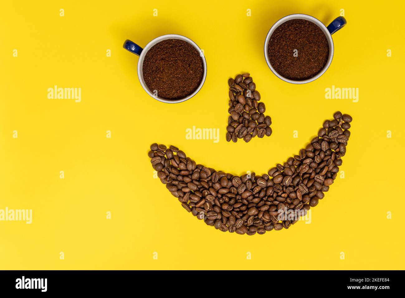Emoticon sorridente, labbra in chicchi di caffè e occhi in tazze di caffè, composizione su sfondo giallo. Foto Stock