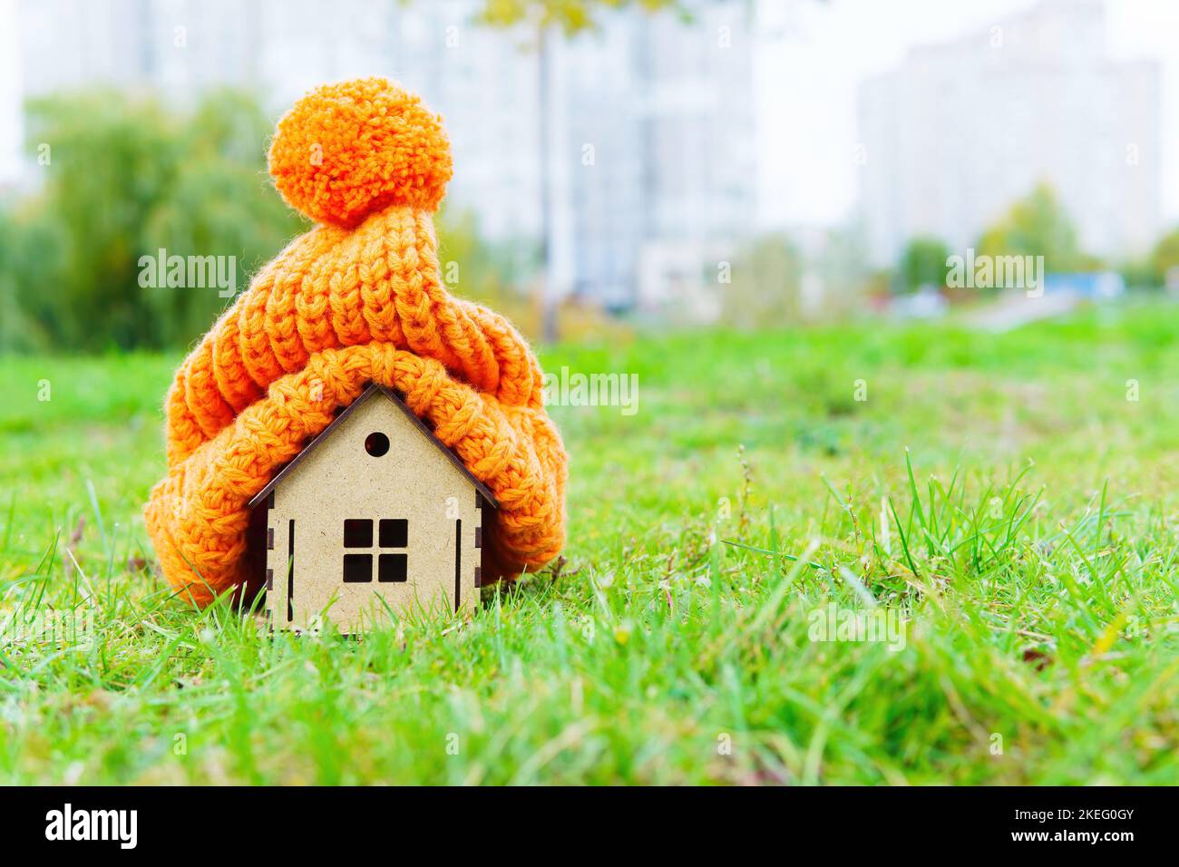 Cappello pompon chunky messo su una statuina di casa di legno disposto su un prato verde all'aperto. Concetto di riscaldamento della casa cost-friendly. Foto Stock