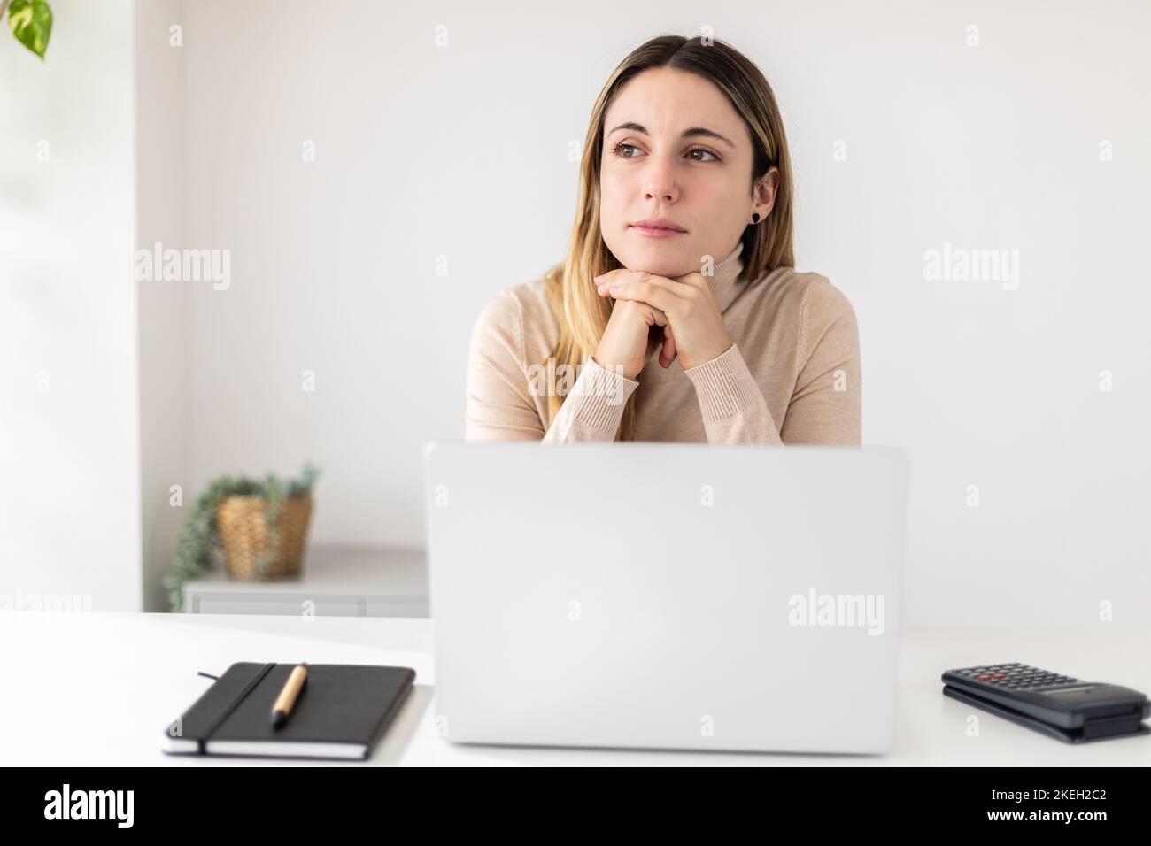 Giovane donna pensierosa e graziosa con un computer portatile seduto alla scrivania che guarda lontano Foto Stock