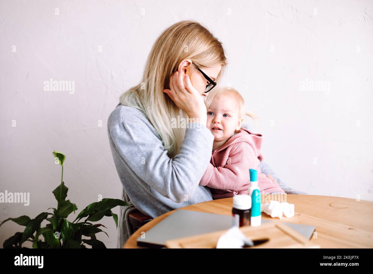 Vista laterale della giovane madre donna seduta a tavolo, tenendo piccola ragazza tristissima bambino, calmando giù a casa. Tempo di malattia. Foto Stock