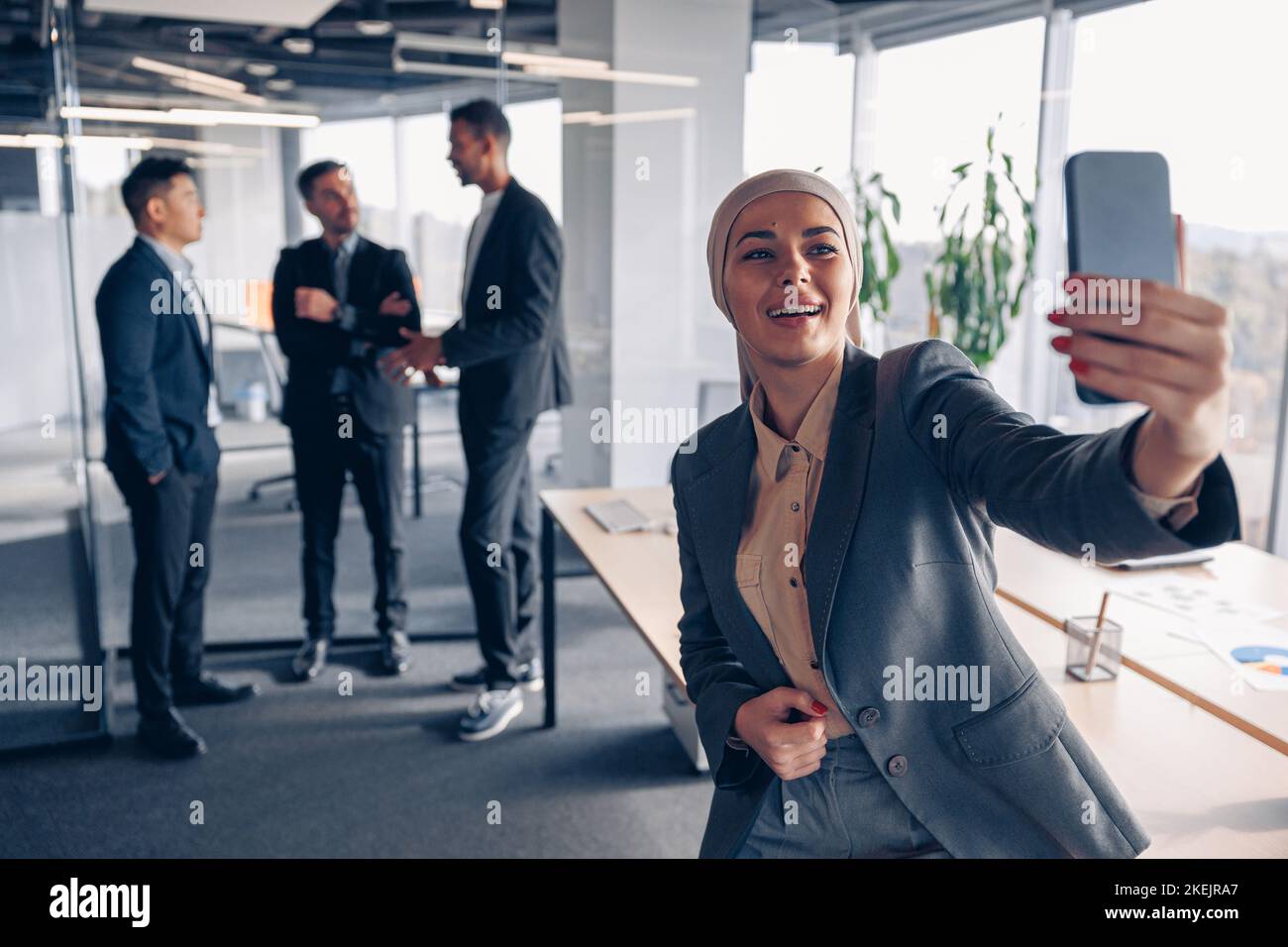 Donna d'affari musulmana sorridente in hijab prendendo un selfie sullo sfondo dei colleghi in ufficio Foto Stock
