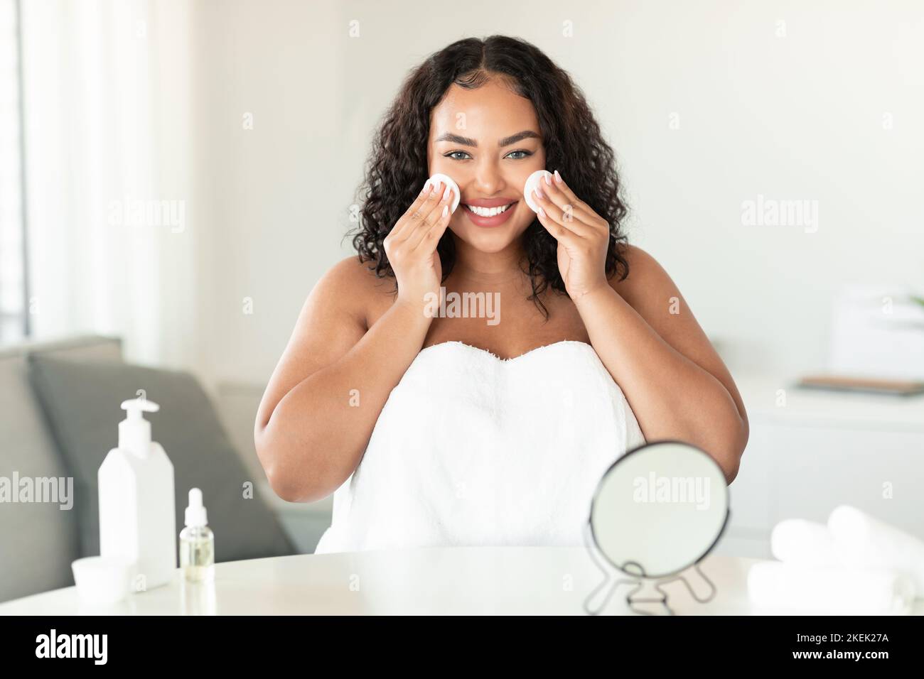 Concetto di cura della pelle. Felice nero più taglia donna utilizzando due tamponi di cotone per la pulizia della pelle, in piedi avvolto in un asciugamano Foto Stock