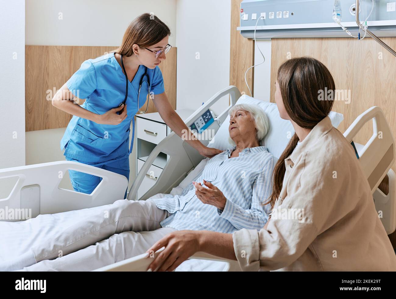 Il parente sostiene la paziente femminile anziana di malattia dell'ospedale sdraiato nel reparto mentre rotondo dell'infermiera del reparto. Assistenza infermieristica Foto Stock