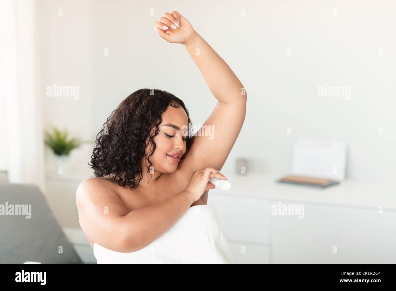 Giovane donna con corpo nero positivo che usa un bastone antitraspirante per le ascelle, seduta in camera da letto, spazio libero Foto Stock