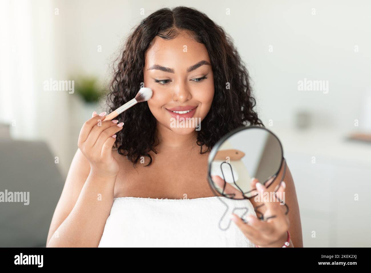 Giovane donna nera corpo positivo che tiene lo specchio e l'applicazione di arrossamento sul viso con il pennello trucco, facendo il trucco quotidiano Foto Stock