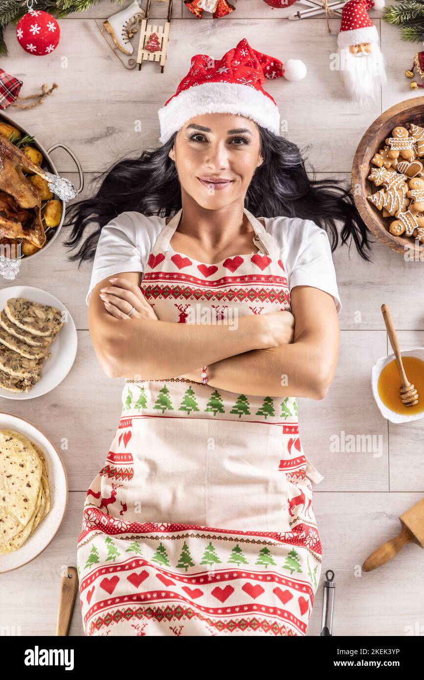 Cuoca femminile soddisfatta dai capelli scuri in un grembiule di Natale e cappello di Santa, sdraiata sul pavimento, circondata da pan di zenzero, dolci Linz, dall'altro lato: roa Foto Stock